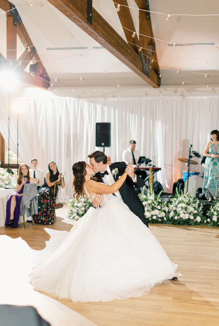 Bride and groom first dance in rustic wedding venue with exposed beams and greenery wall