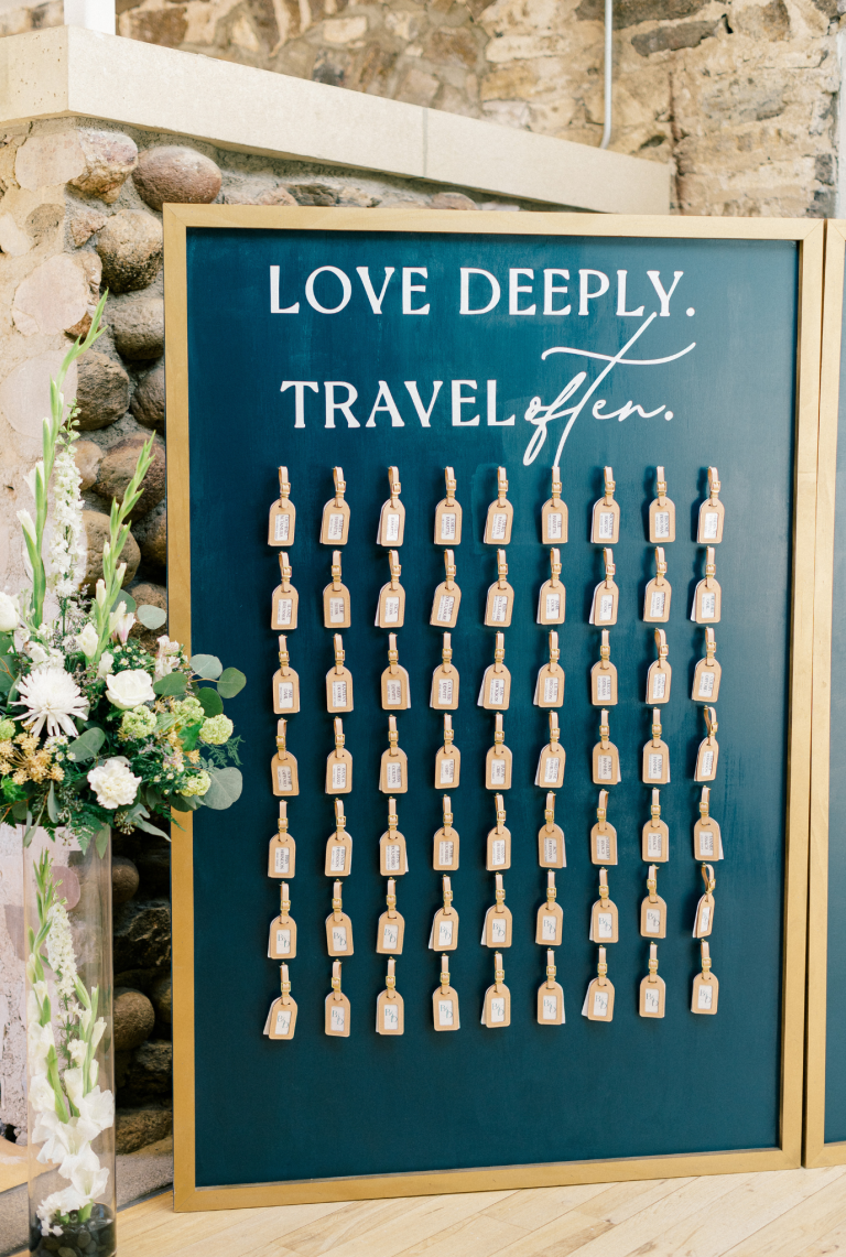 Travel-themed wedding seating chart with wooden luggage tags arranged on teal board reading "Love Deeply. Travel Often."