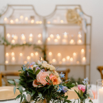 Peach rose and greenery centerpiece on white table with illuminated candle shelves in background