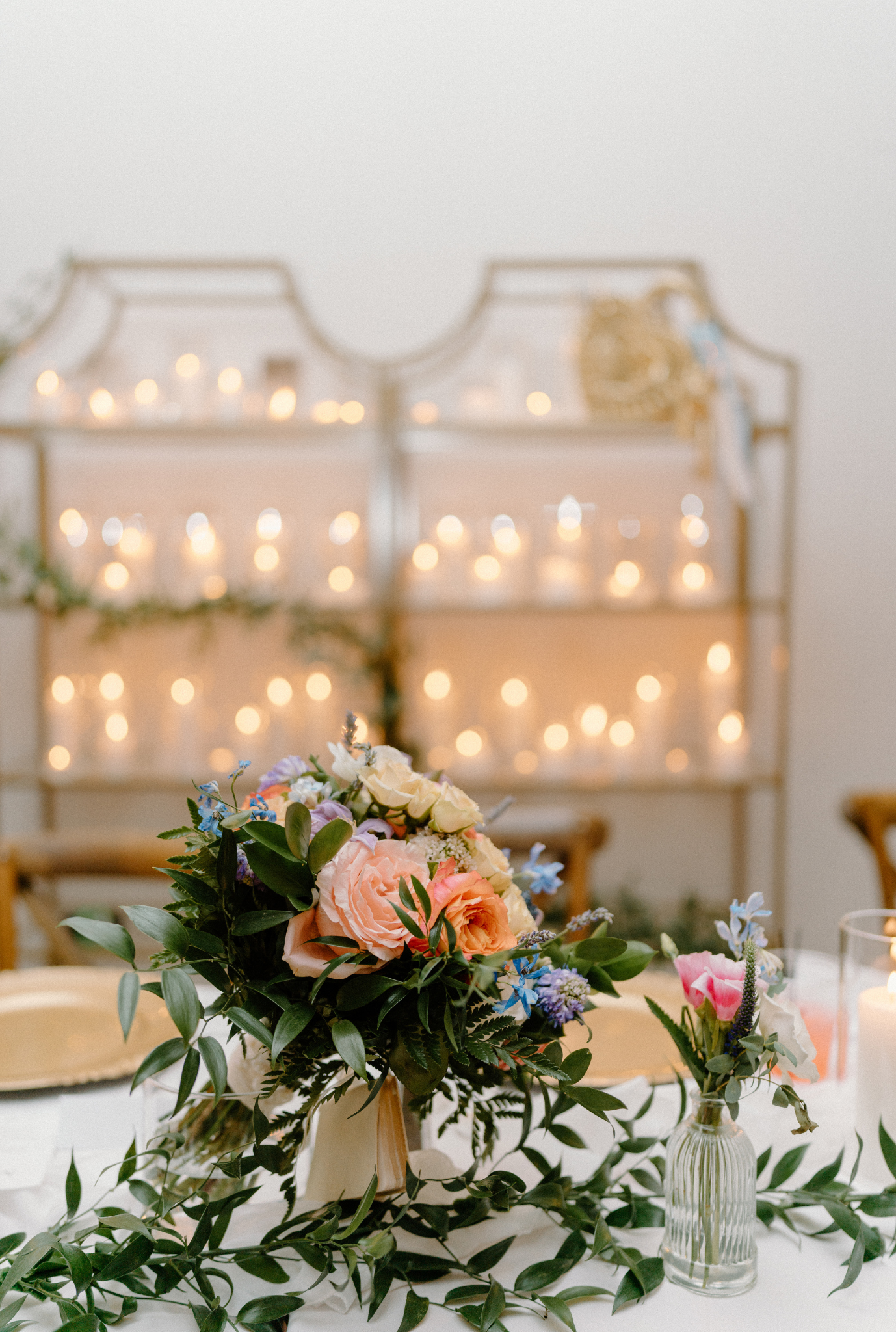 Peach rose and greenery centerpiece on white table with illuminated candle shelves in background