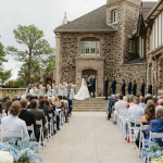 Outdoor wedding ceremony at stone castle venue with guests seated facing wedding party on steps