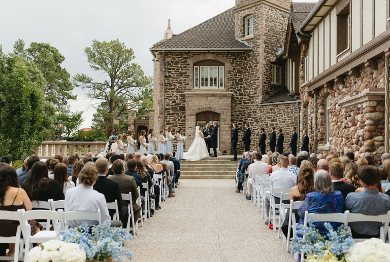 Outdoor wedding ceremony at stone castle venue with guests seated facing wedding party on steps
