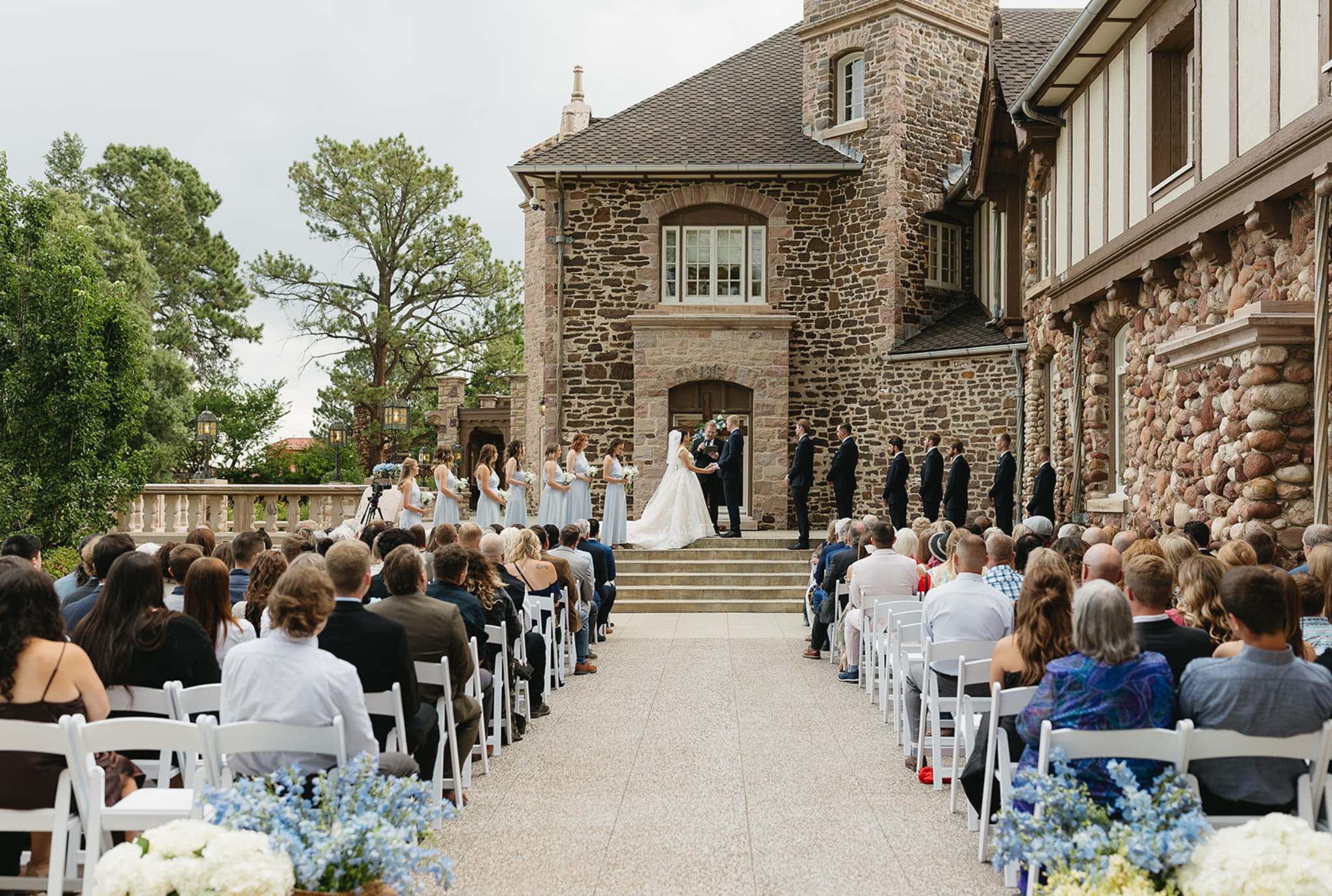 Outdoor wedding ceremony at stone castle venue with guests seated facing wedding party on steps