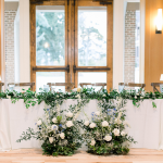 Elegant wedding sweetheart table with lush greenery garland and white floral arrangements against bright windows