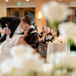 Bride and groom sharing romantic dip kiss during reception as guests watch from head table