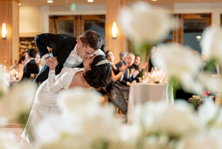 Bride and groom sharing romantic dip kiss during reception as guests watch from head table