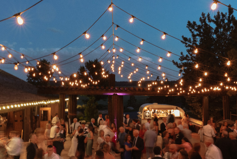 Outdoor wedding reception at dusk with string lights canopy and guests dancing on patio