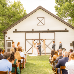 Outdoor barn wedding ceremony with guests seated on lawn facing white barn venue