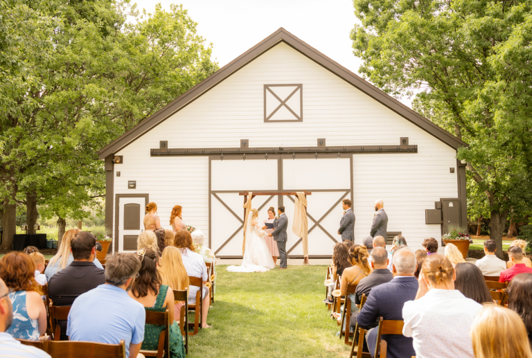 Outdoor barn wedding ceremony with guests seated on lawn facing white barn venue