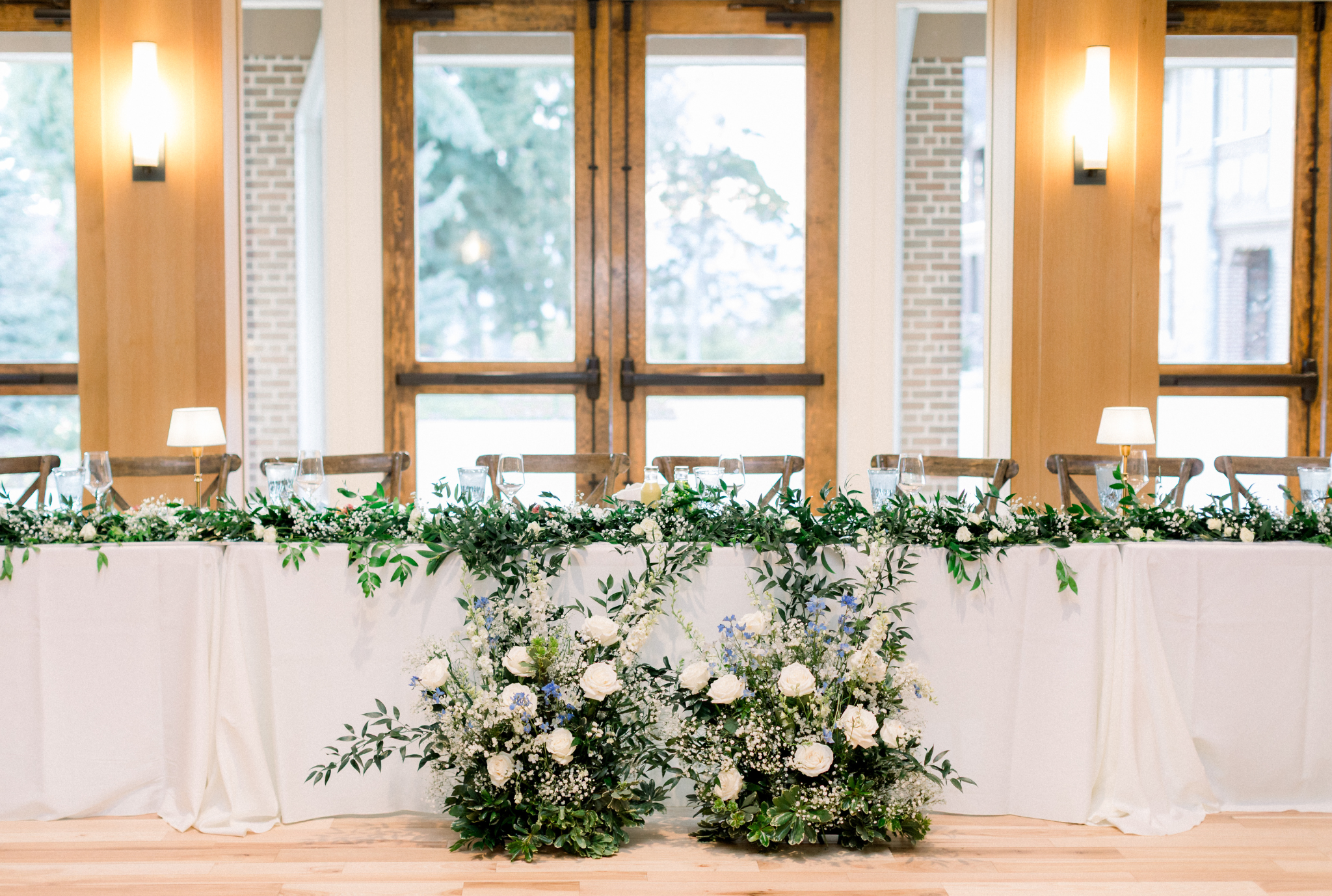 Elegant wedding sweetheart table with lush greenery garland and white floral arrangements against bright windows