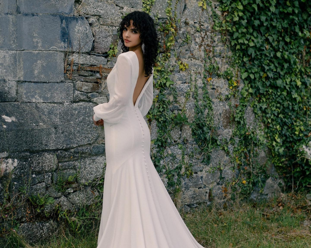 Woman in elegant long-sleeved blush wedding gown with open back and mermaid silhouette against ivy-covered stone wall