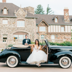Bride and groom posing with vintage black convertible car in front of stone mansion venue in Denver