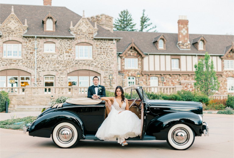 Bride and groom posing with vintage black convertible car in front of stone mansion venue in Denver