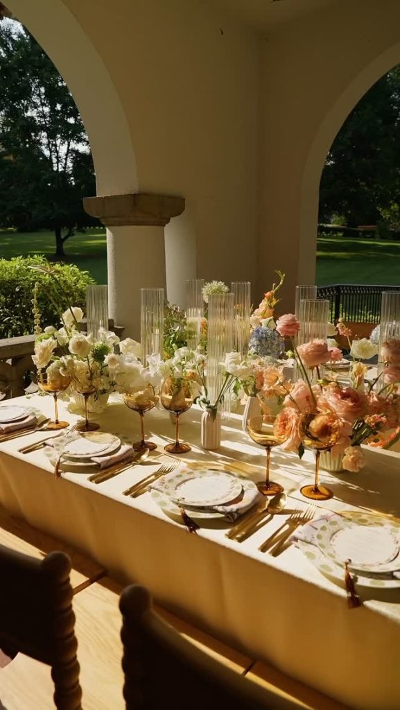 Elegant outdoor wedding reception table with gold stemmed floral centerpieces, white place settings, and glass cylinder vases under covered archway