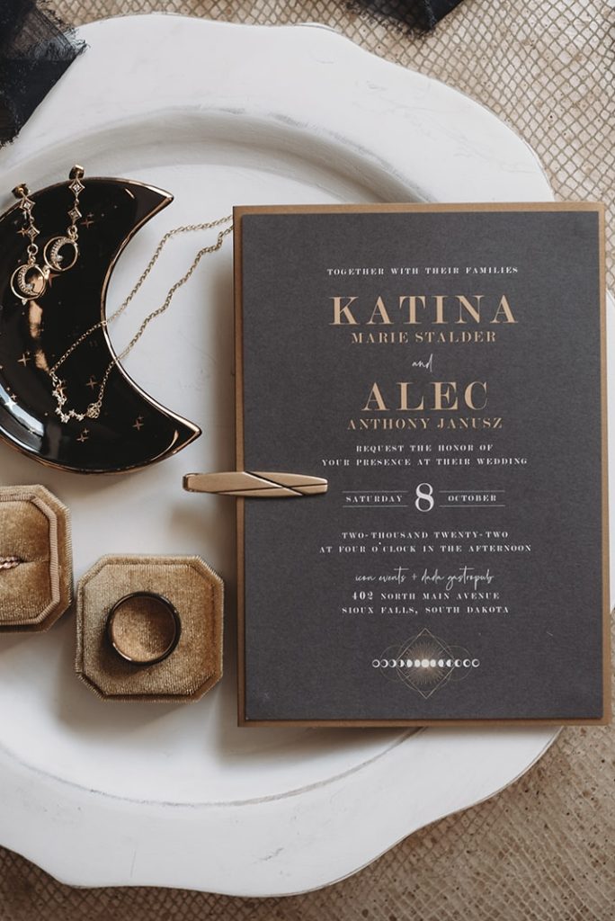 Dark gray and gold wedding invitation with modern typography displayed on white plate alongside wedding rings and moon-themed jewelry