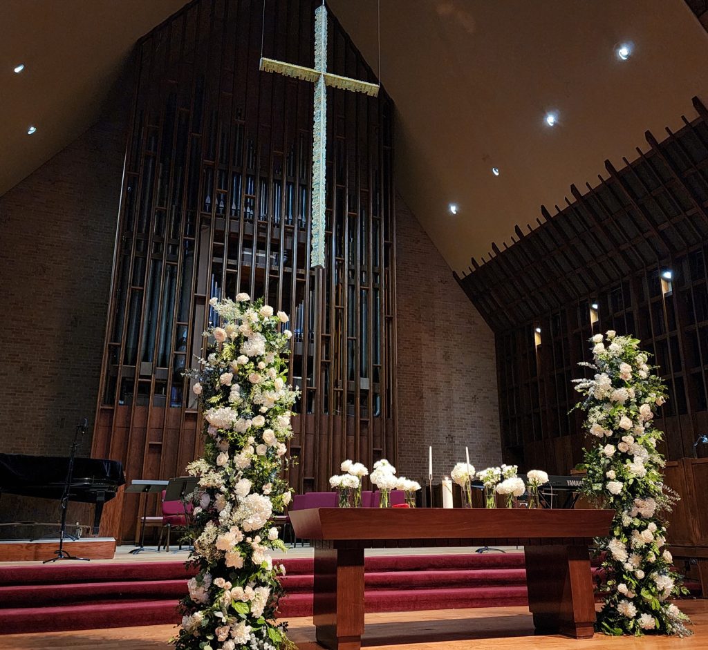 Church sanctuary altar decorated with tall white and blush floral arrangements flanking wooden communion table beneath large cross and pipe organ