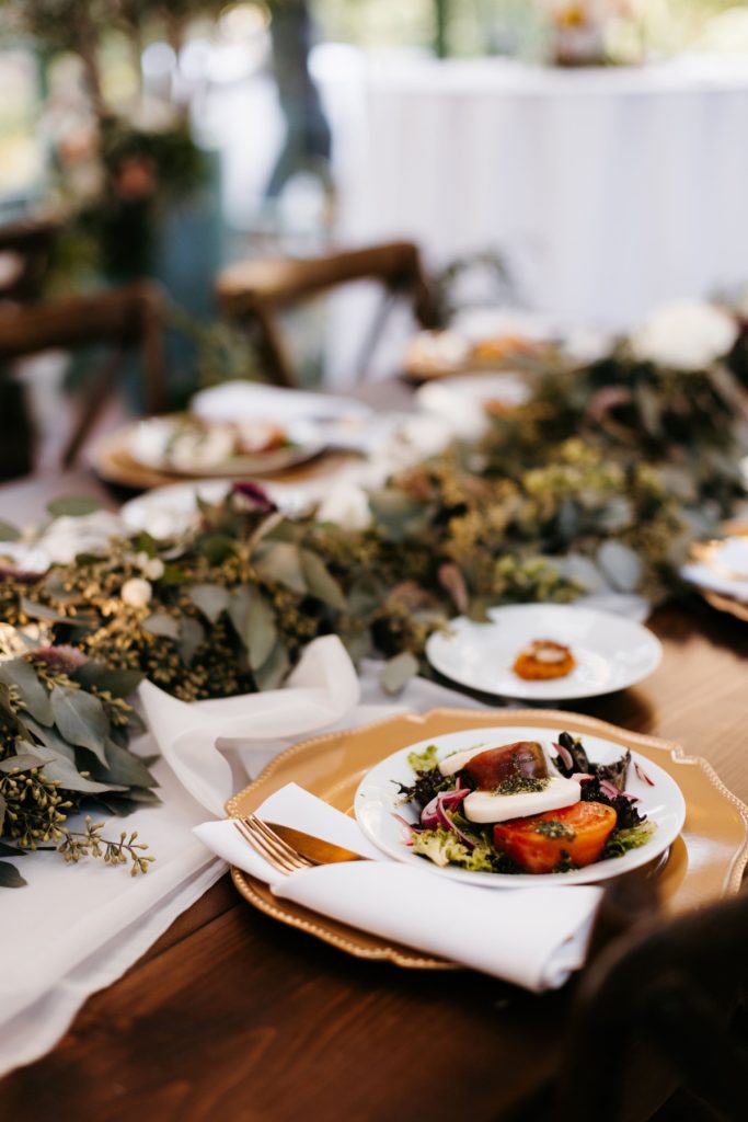Elegant wedding table setting with heirloom tomato salad on gold charger, eucalyptus garland centerpiece, and wooden chairs
