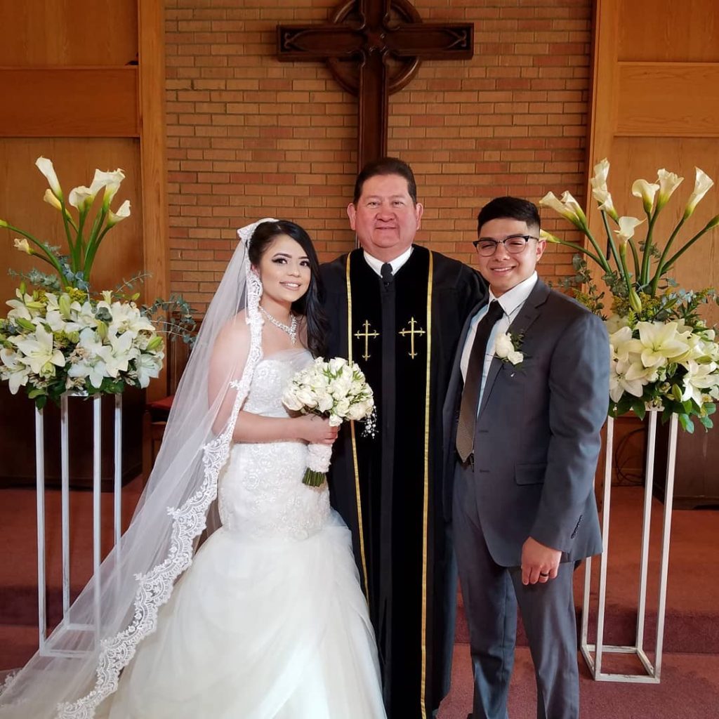 Bride and groom with wedding officiant in church sanctuary decorated with white floral arrangements