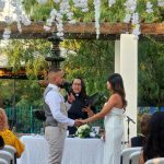 Outdoor wedding ceremony with officiant leading couple through vows under wooden pergola decorated with white flowers