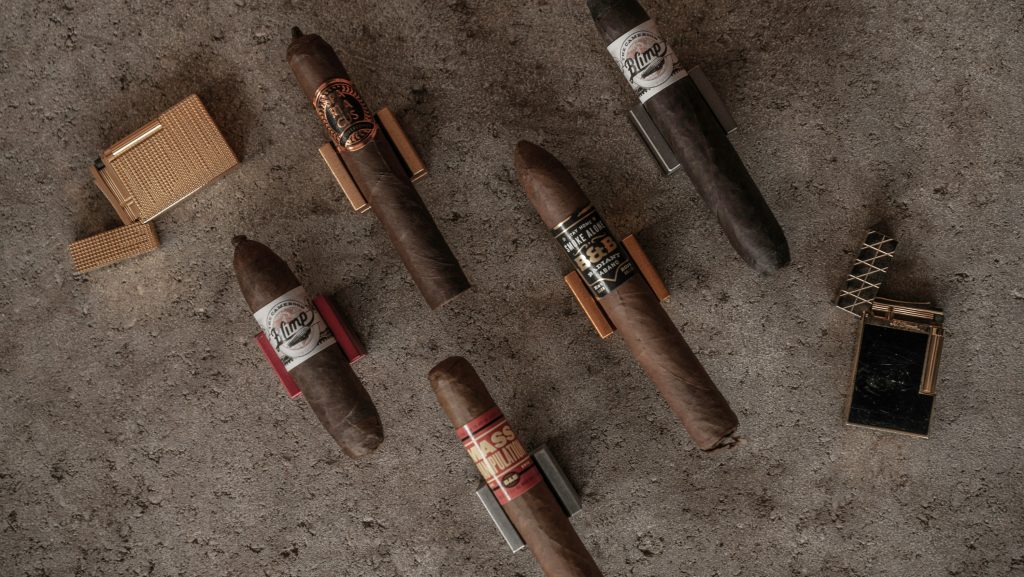 Assortment of premium cigars with decorative bands displayed alongside elegant gold lighters on concrete surface