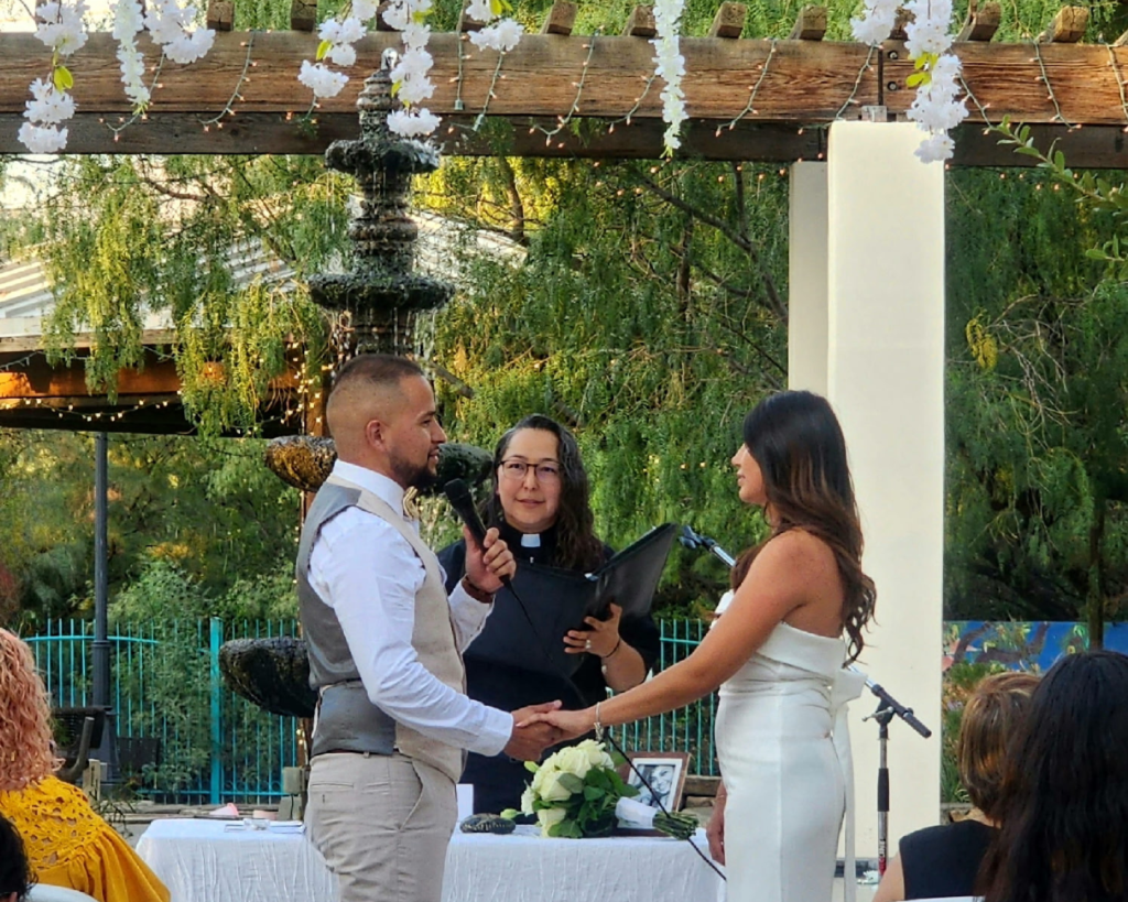 Wedding officiant conducting outdoor ceremony for couple holding hands under pergola decorated with white flowers and string lights