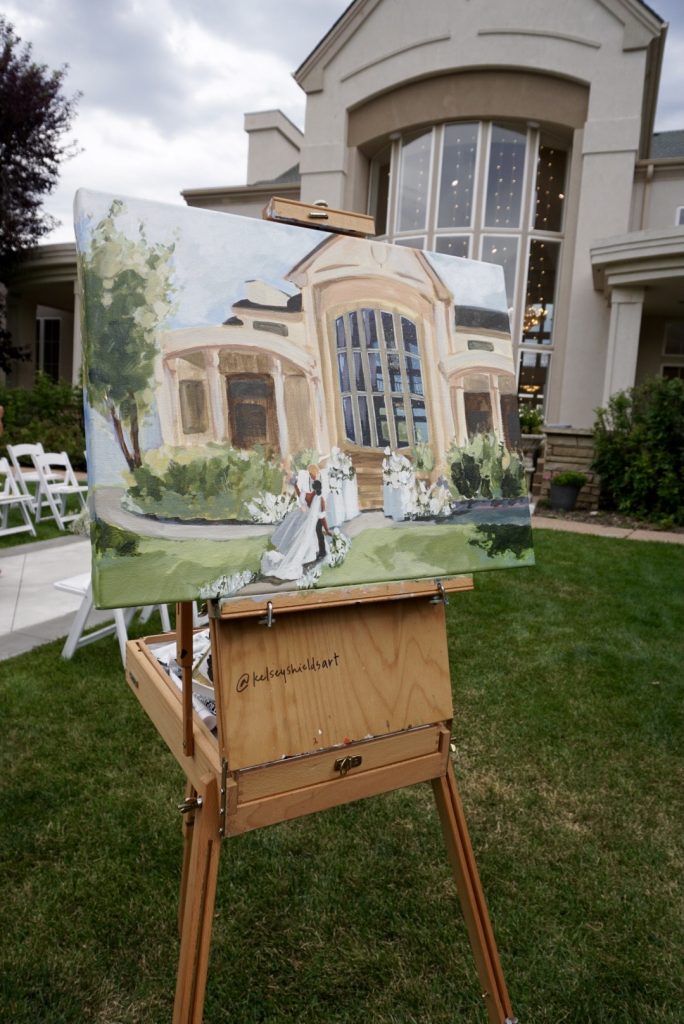 Live wedding painting on easel showing elegant mansion venue with bride walking through white floral arrangements