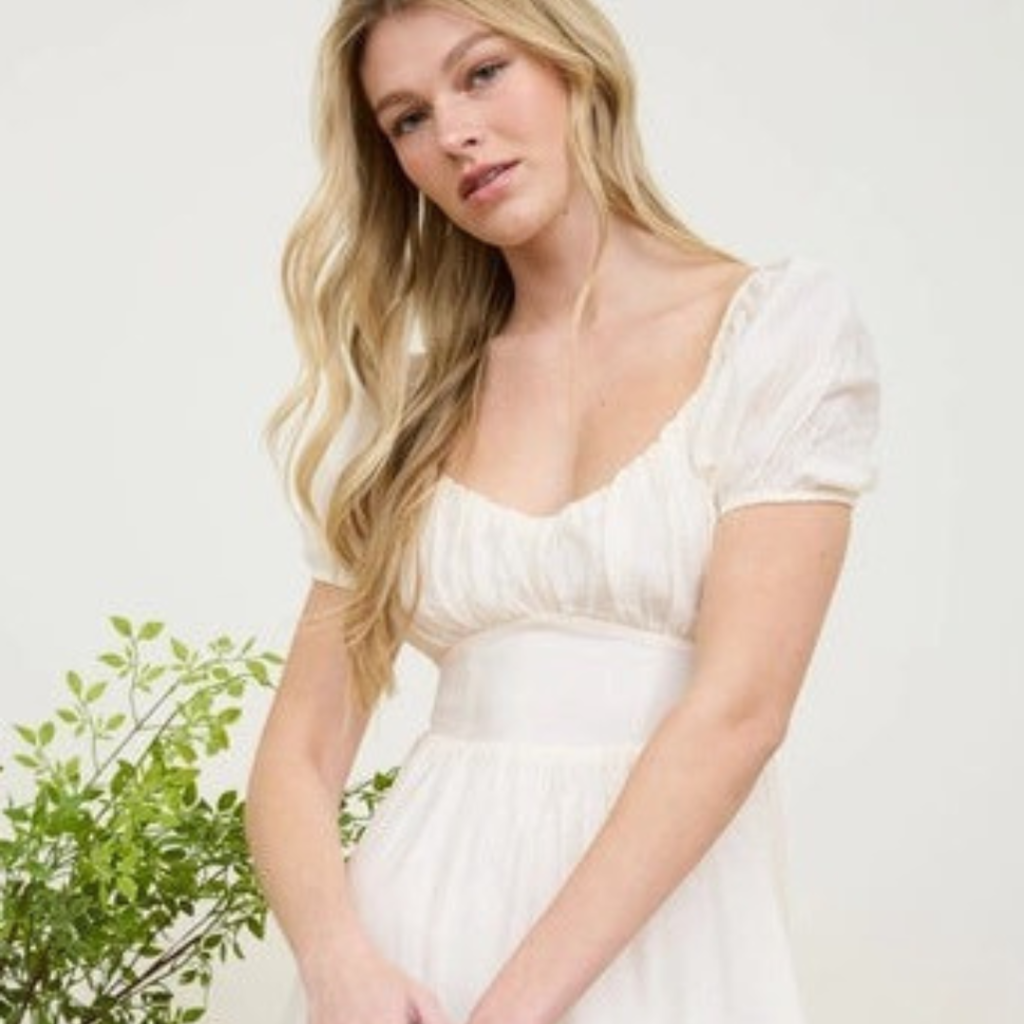 Blonde woman in white empire-waist dress with puff sleeves holds greenery and white flowers against neutral background