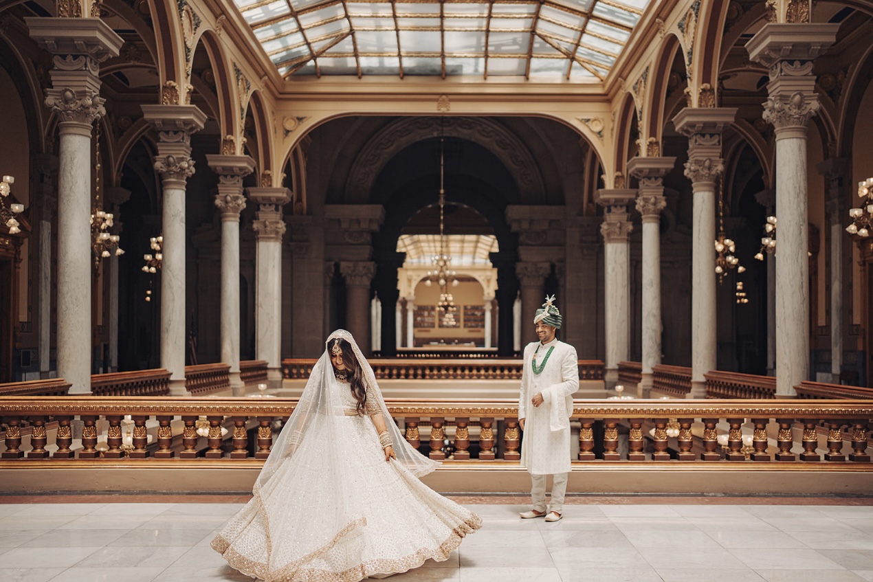 Bride and groom in traditional Indian wedding attire pose in ornate columned atrium with skylight