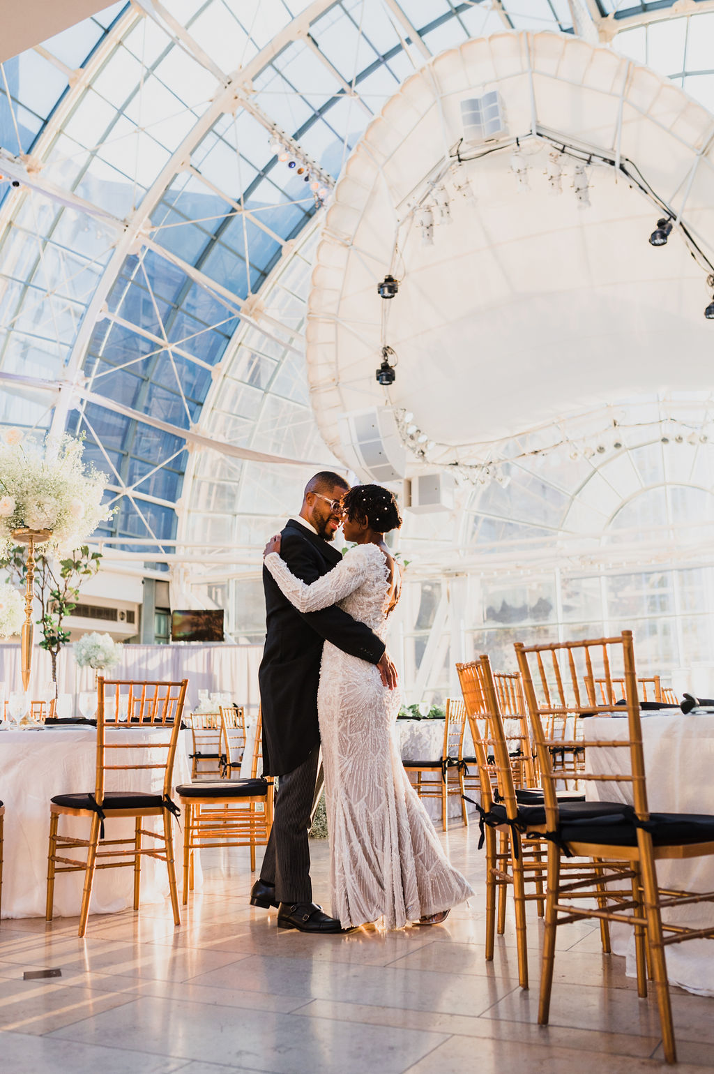 Couple sharing first dance under dramatic glass atrium ceiling with gold chiavari chairs