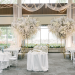 Elegant white wedding reception with tall white floral centerpieces, draped fabric, and string lights