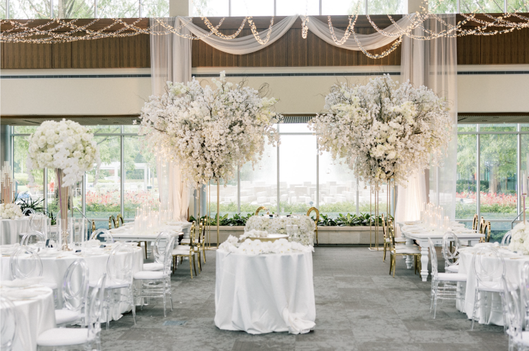 Elegant white wedding reception with tall white floral centerpieces, draped fabric, and string lights