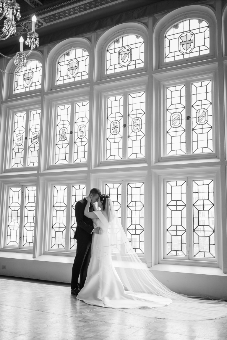 Bride and groom embracing before ornate arched leaded glass windows with crystal chandelier