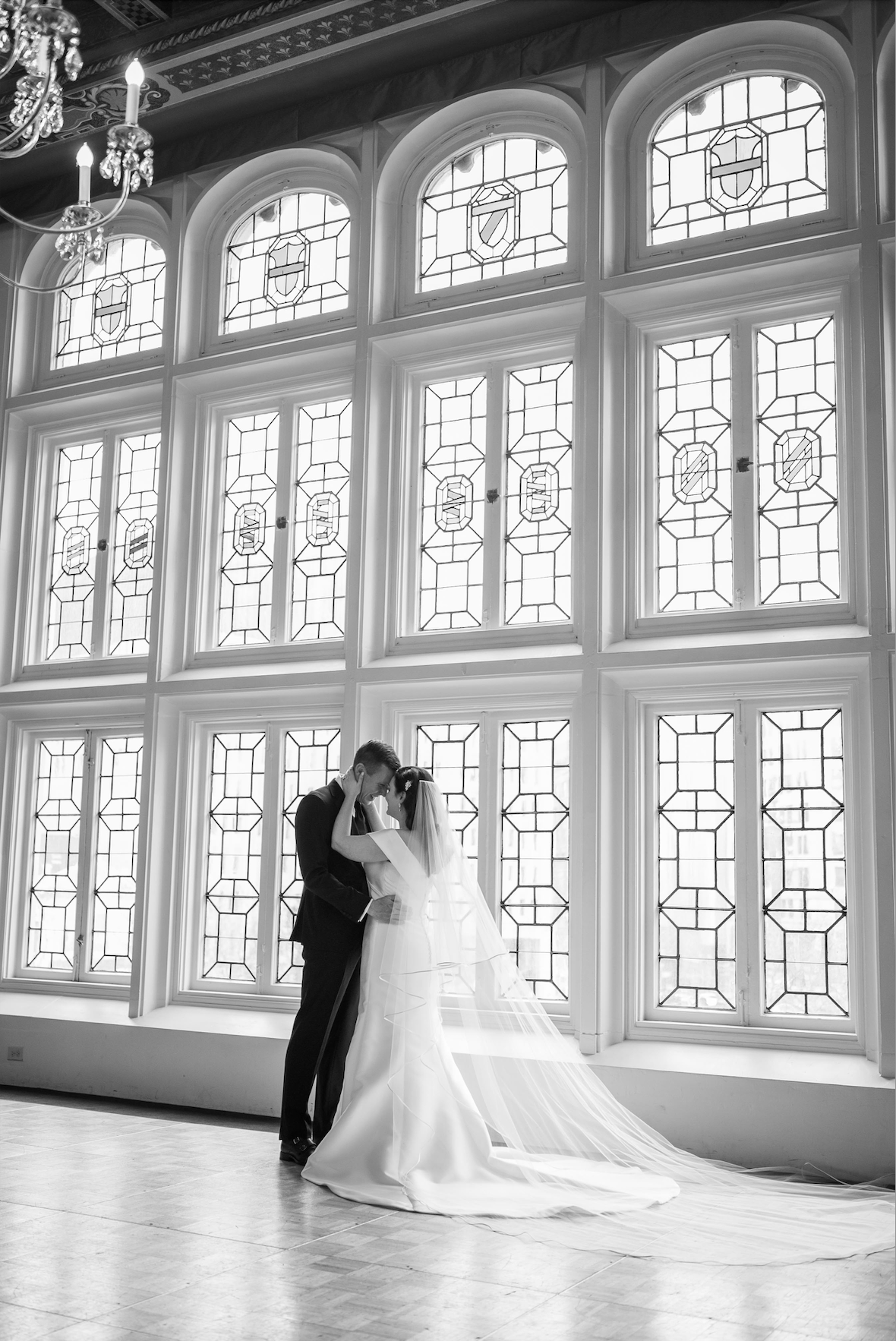 Bride and groom embracing before ornate arched leaded glass windows with crystal chandelier
