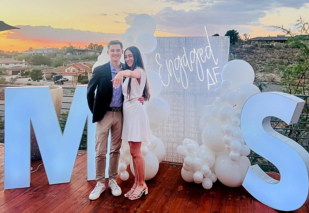 Newly engaged couple celebrating at rooftop proposal with large "M + S" letters, white balloons, and "Engaged AF" neon sign at sunset