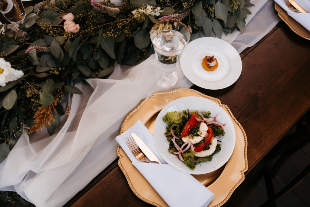 Elegant plated wedding meal with greens and garnish on wooden charger beside wine glass