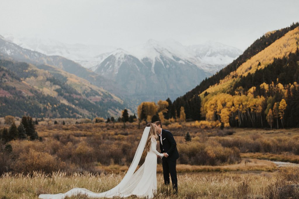 Bride and groom kissing in autumn mountain valley with golden aspens and snow-capped Colorado peaks