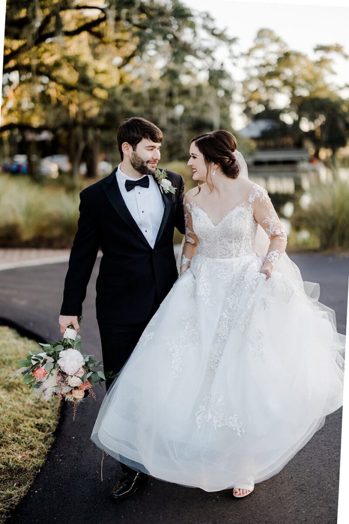 Bride in lace ballgown and groom in tuxedo walking together beneath oak trees