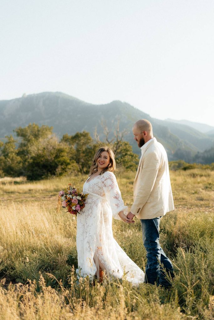 Bride and groom hold hands in sunlit mountain field at golden hour