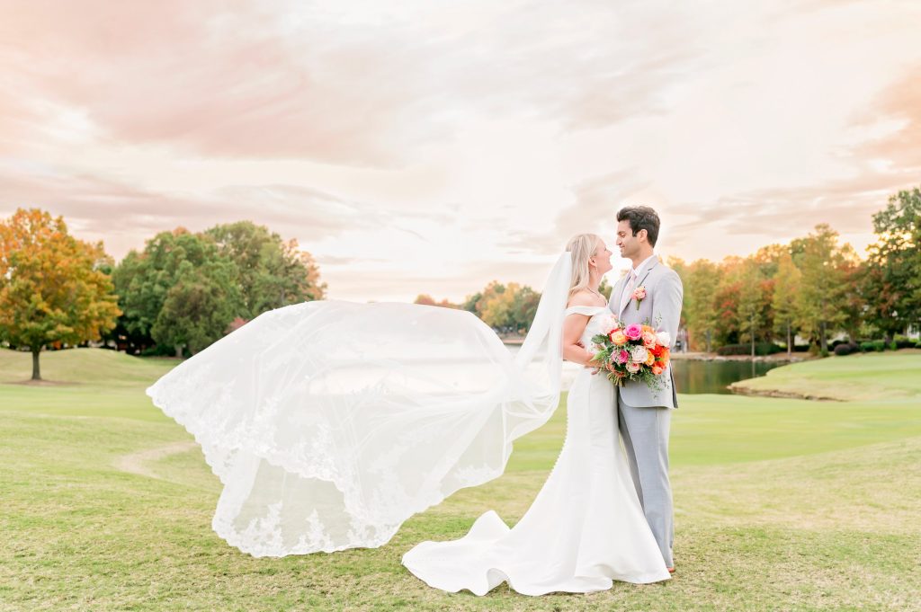 Bride's veil billowing in wind as couple embraces on golf course at sunset with colorful bouquet
