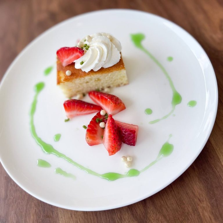 Plated tres leches cake dessert with whipped cream, fresh strawberry garnish, and decorative green sauce drizzle