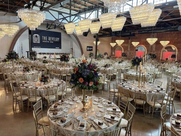 The Revelry North End industrial reception venue with crystal chandeliers, gold chiavari chairs, and tall floral centerpieces