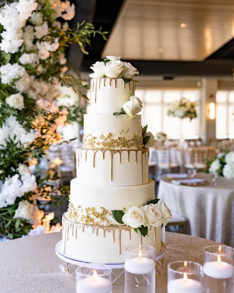 Five-tier white wedding cake with gold drip detail, fresh white roses, gold leaf accents, and surrounded by votive candles