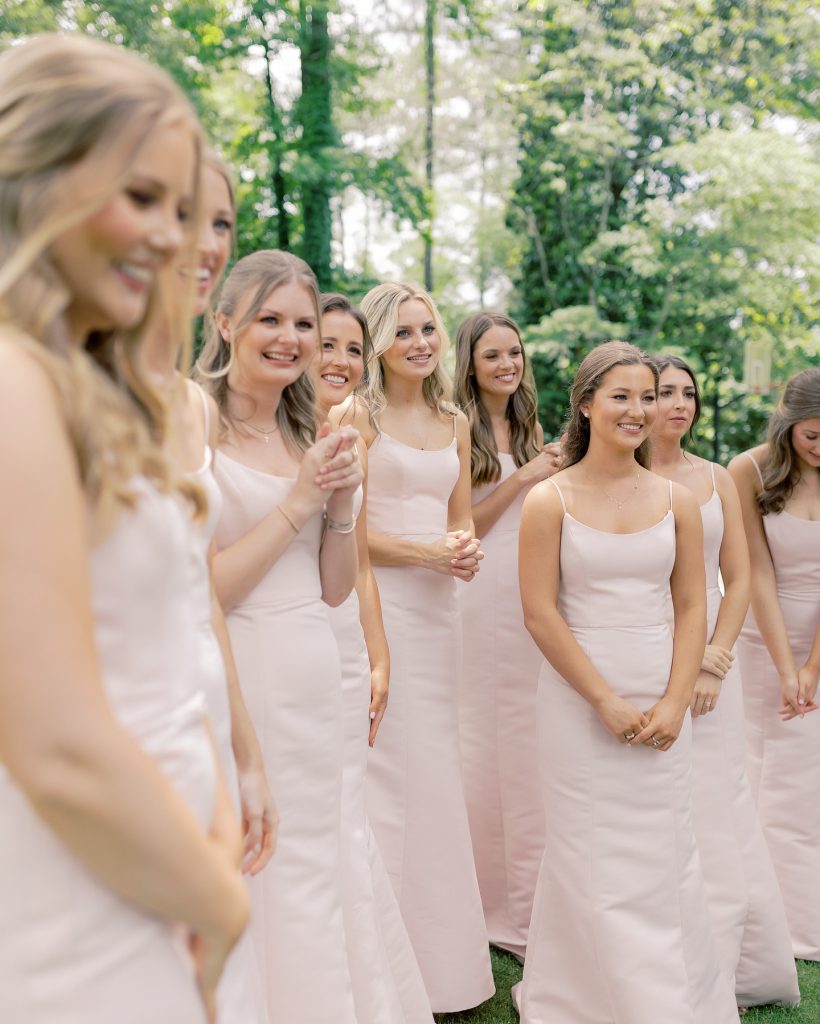 Bridesmaids in matching blush pink dresses lined up outdoors among lush green trees