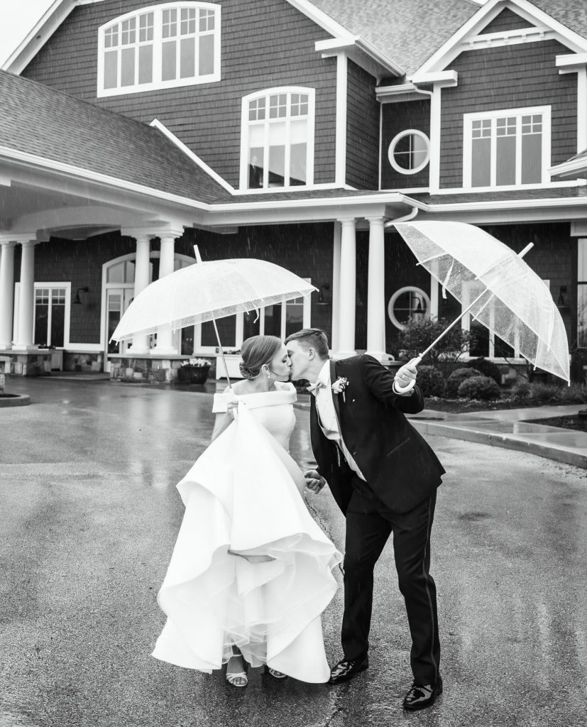 Bride and groom kissing under umbrellas in the rain outside Chatham Hills clubhouse
