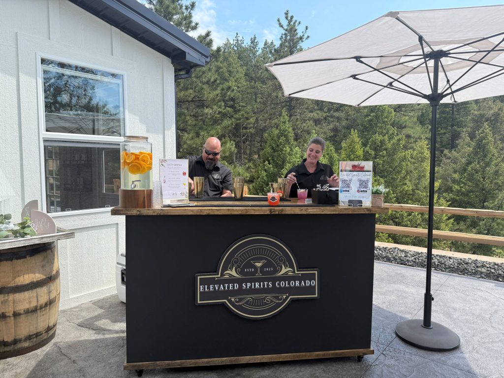 Two bartenders preparing cocktails at outdoor mobile bar under white umbrella with mountain forest backdrop