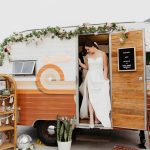 Bride in strapless white gown stepping out of retro orange and white photo booth camper trailer decorated with floral garland