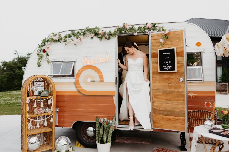 Bride in strapless white gown stepping out of retro orange and white photo booth camper trailer decorated with floral garland