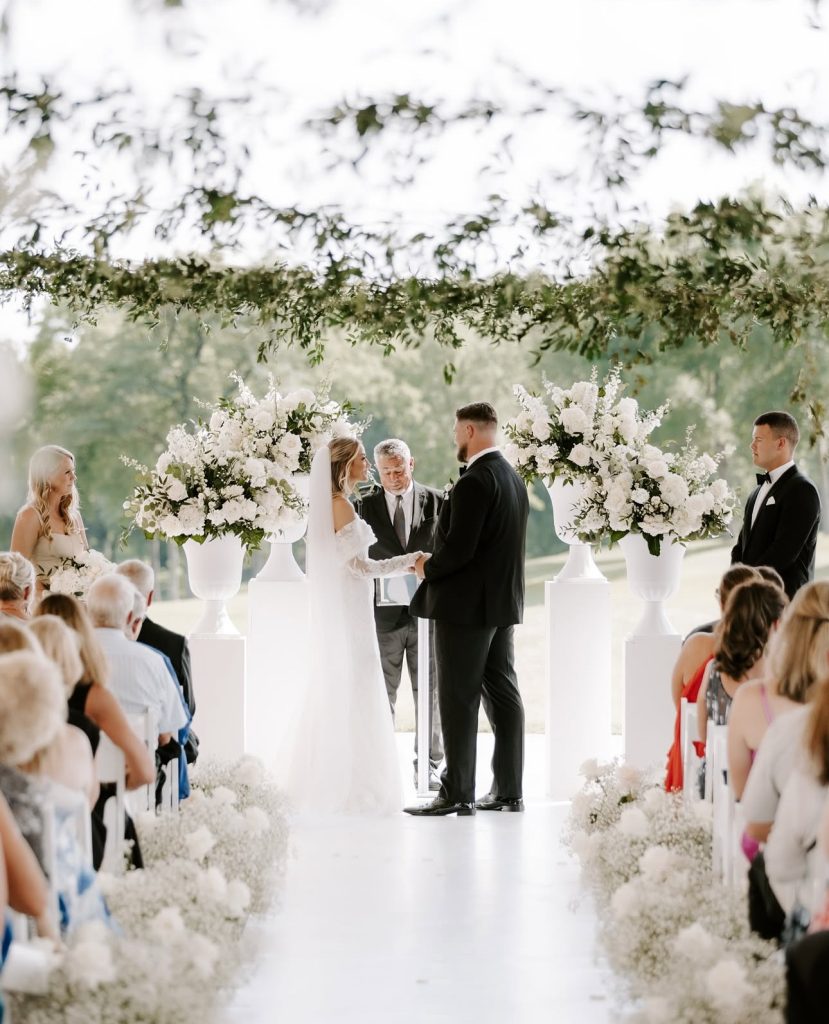 Outdoor ceremony under clear tent with white floral arrangements and greenery garland overhead