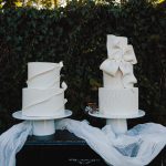 Two elegant white wedding cakes with flowing fondant drapes and bow details displayed on pedestals with dusty blue fabric