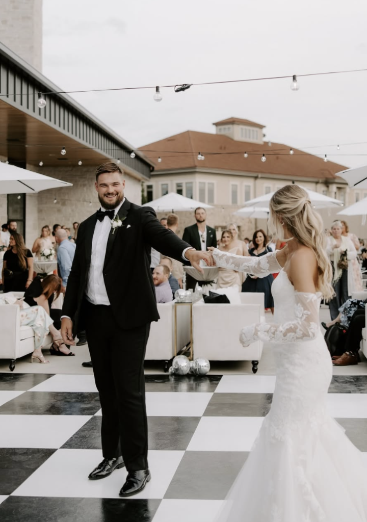 Bride and groom share first dance on checkered floor at outdoor reception with guests watching